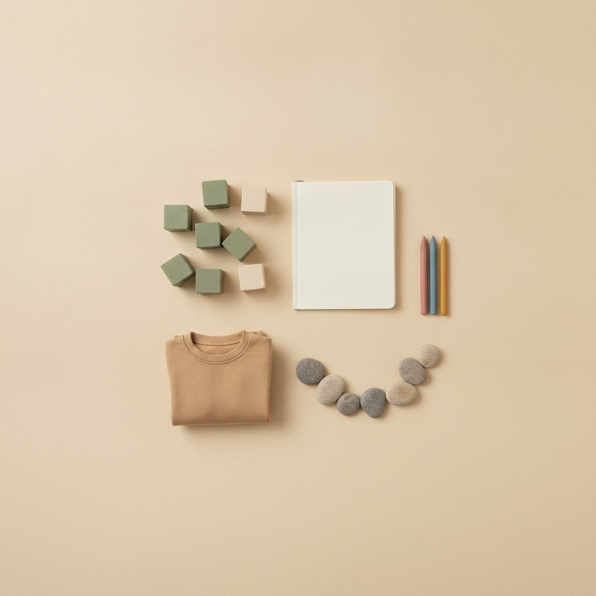 Sage and natural wooden blocks, a blank notebook with three crayons, a curve of pebbles and a folded tan crew-neck top on a sand background